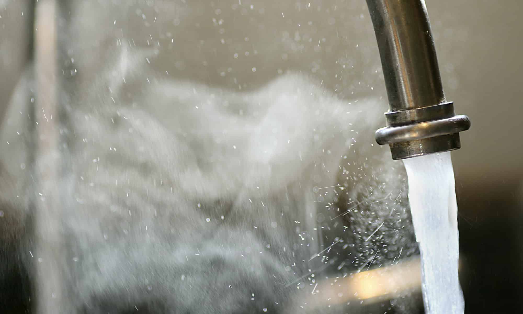 Hot steaming running tap water is pouring out of a stainless steel kitchen faucet.